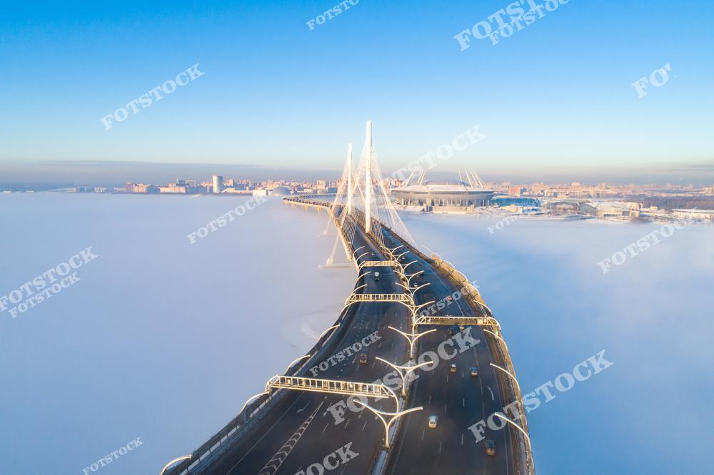 На фотографии запечатлён Большой Обуховский (Вантовый) мост — часть Западного скоростного диаметра, соединяющего районы Санкт-Петербурга. Вид сверху открывает панораму города, окутанного плотным утренним туманом. Мост словно парит над облаками, а на заднем плане виден стадион на Крестовском острове. Яркое зимнее солнце освещает ледяной покров Финского залива. Это один из самых живописных и технологичных мостов города.